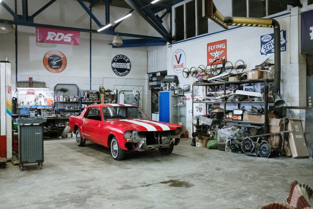 A red classic car under restoration inside a cluttered automotive workshop.