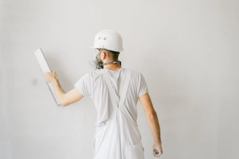 A worker in protective gear smoothing a white wall with a trowel.