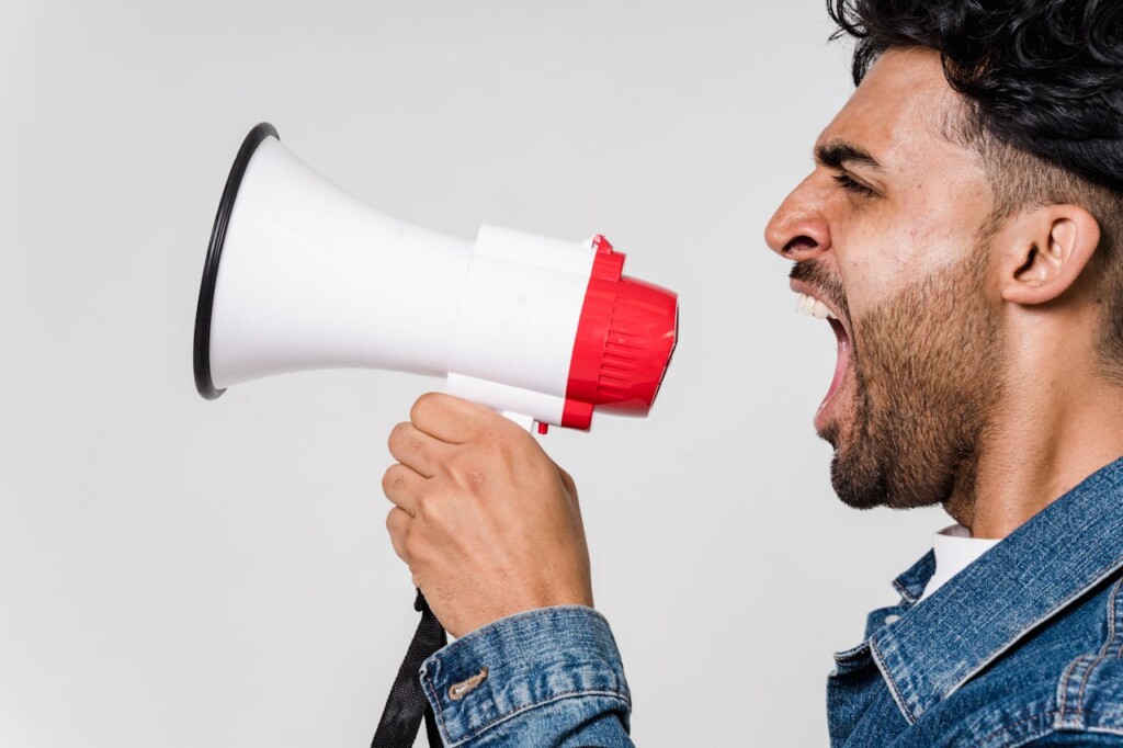 A man in a denim jacket shouting into a red and white megaphone.