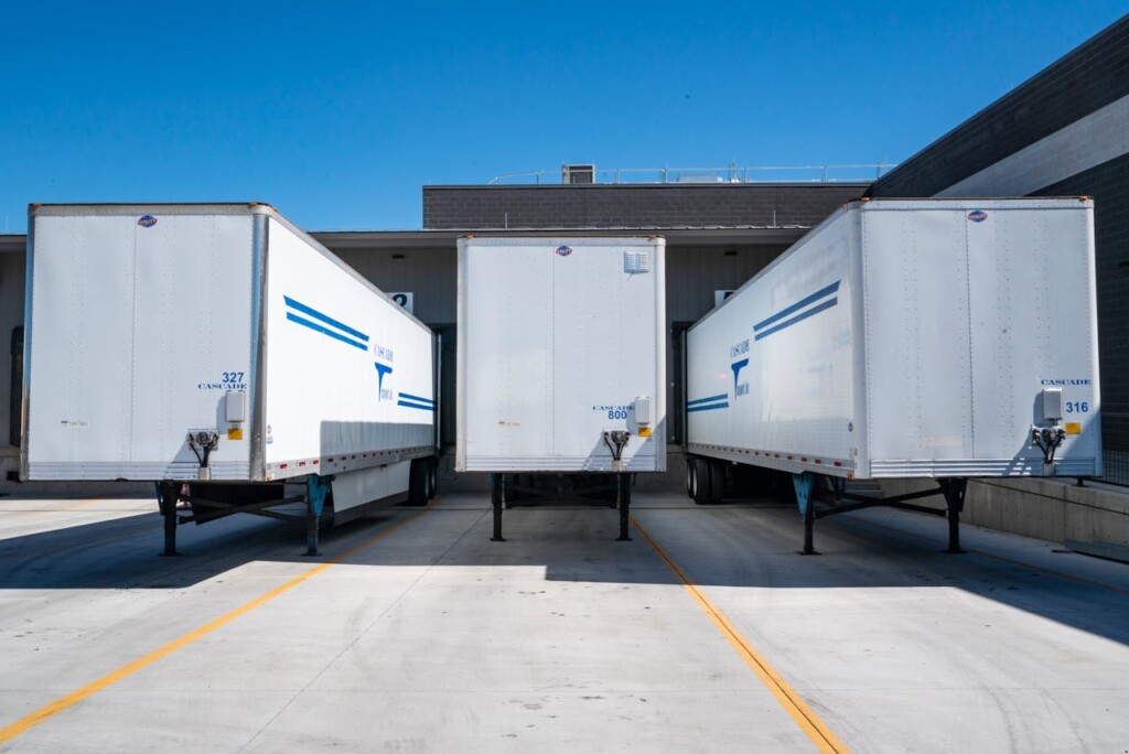 Three white trailers are parked at a loading dock under a clear blue sky.