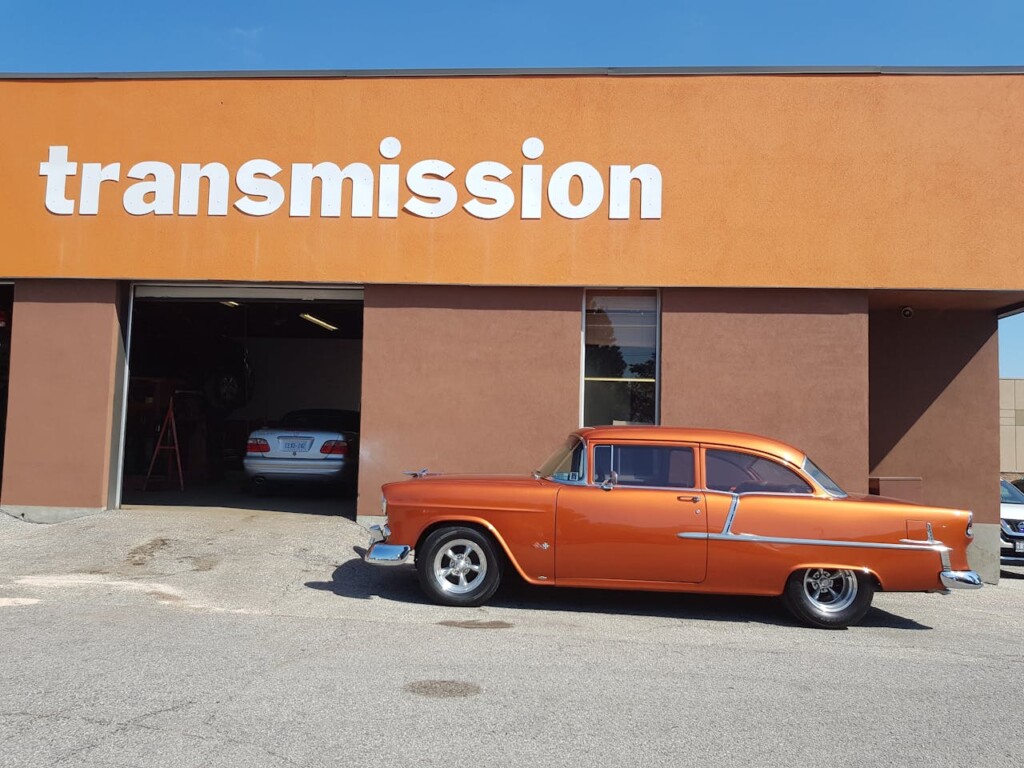 An orange vintage car parked outside an auto repair shop labeled "transmission."