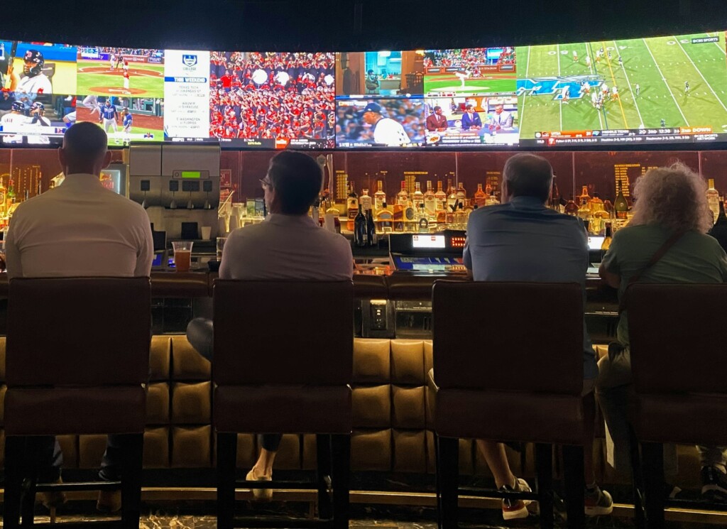 Four people watch multiple sports on large TV screens at a bar.