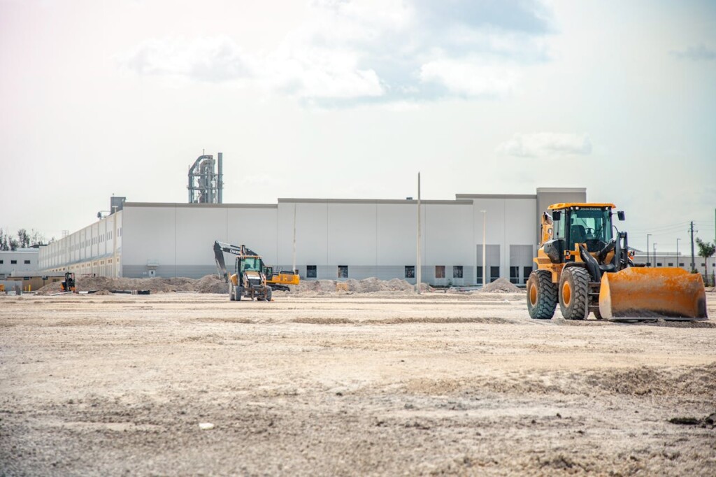 Construction vehicles on a large barren site with a factory in the background.