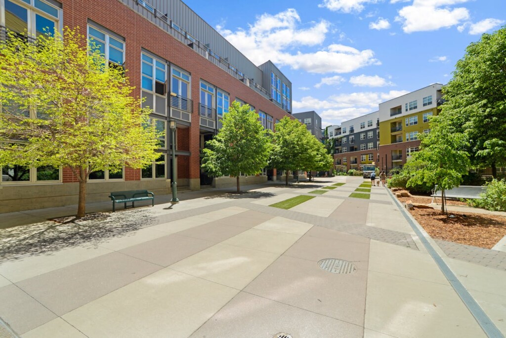 Modern urban walkway with bright trees and multistory buildings under a blue sky.