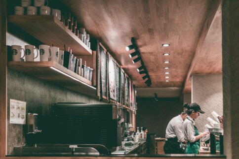 Baristas working behind a coffee shop counter with menus and shelves nearby.