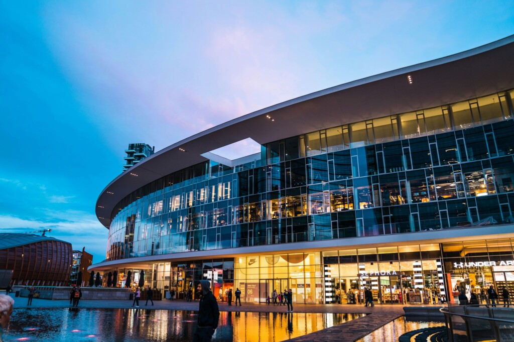 Modern shopping mall with glass facade reflecting evening lights and people walking.