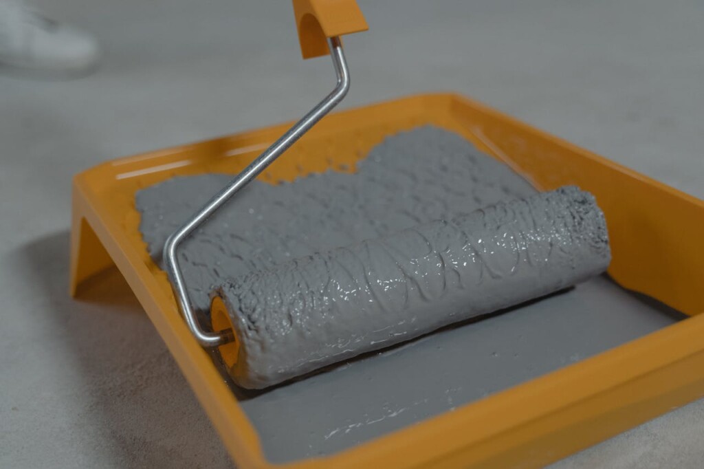 A paint roller in a tray with gray paint on a concrete floor.