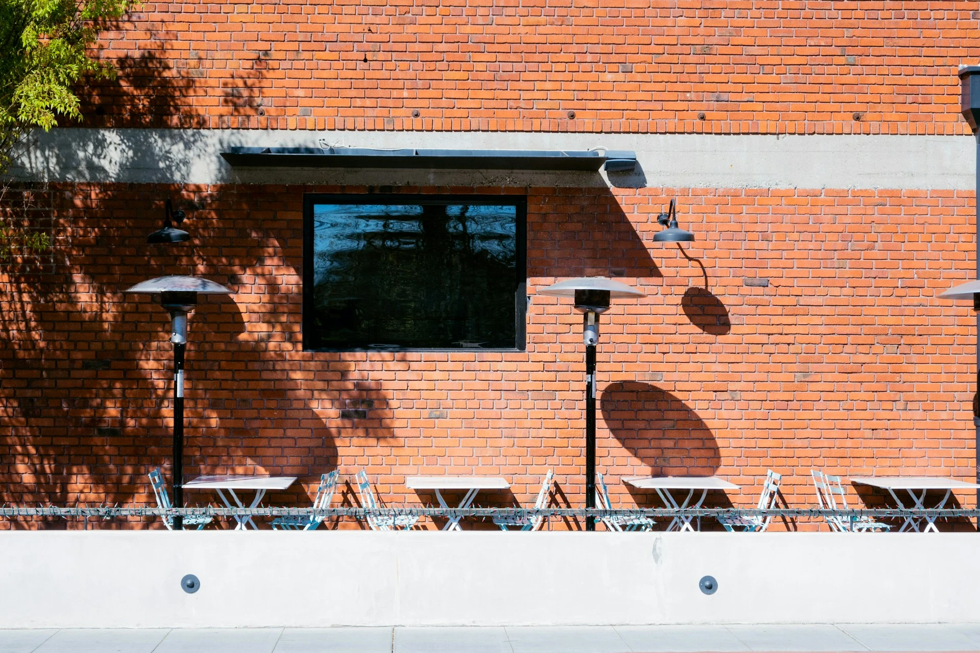 Outdoor cafe seating against a red brick wall with heaters and shadows.